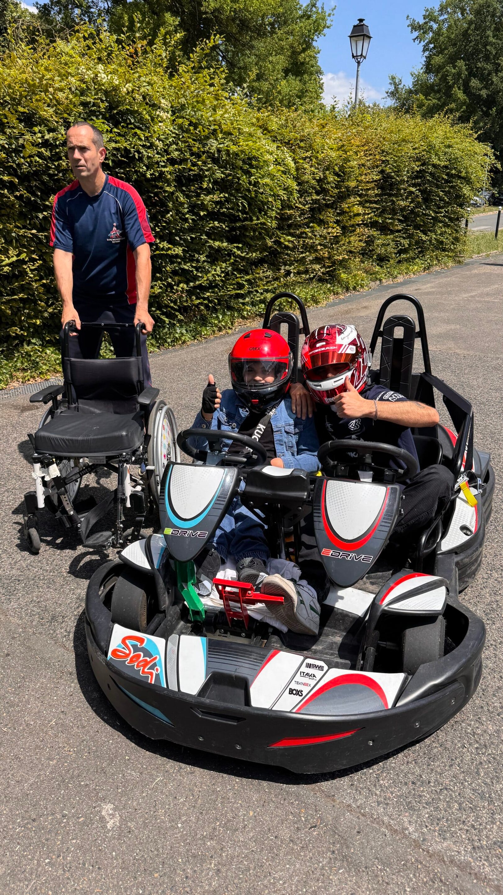 Deux personnes en kart, dont un enfant porteur de handicap sourient et font signe.