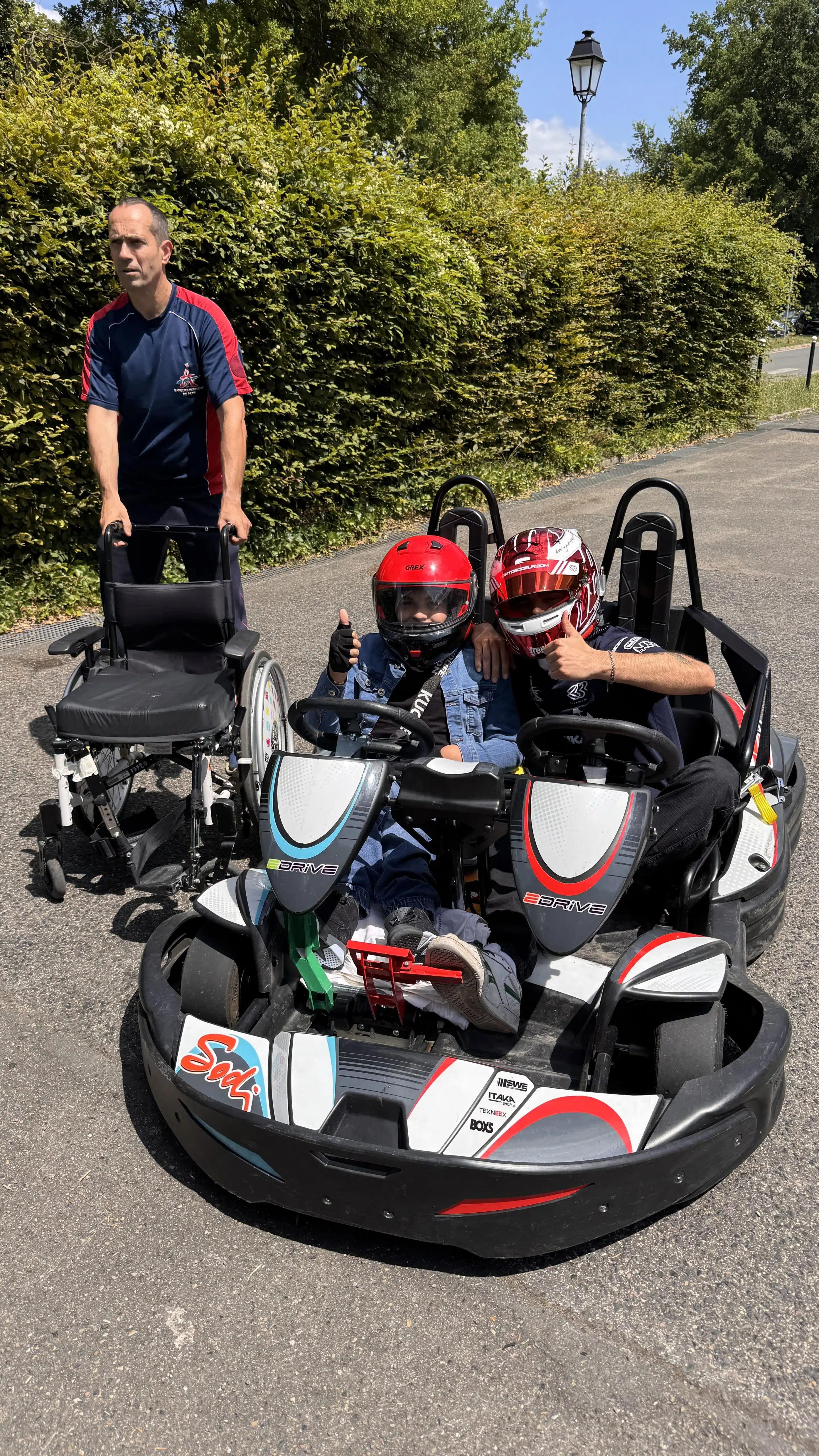 Deux personnes en kart, dont un enfant porteur de handicap sourient et font signe.